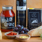 Assorted food items including a jar of Tony Mandina's Red Gravy  bottle of wine, olive oil, grapes, and bread on a wooden surface.